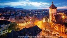 rechts ein altes Rathaus mit Turm, daneben erstreckt sich eine Stadt. Es dämmert und die Straßen sind vom gelben Licht der Laternen erleuchtet.