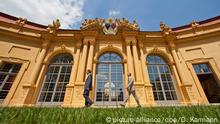 ein Teil der Fassade eines gelbes Barockgebäudes, davor zwei Männer, die nach links gehen (Erlangen Orangerie)