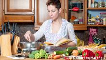 Eine Frau steht in einer Küche, hält ein Kochbuch in der linken Hand und rührt mit der rechten in einem Topf. Vor ihr liegen unterschiedliche Gemüse.