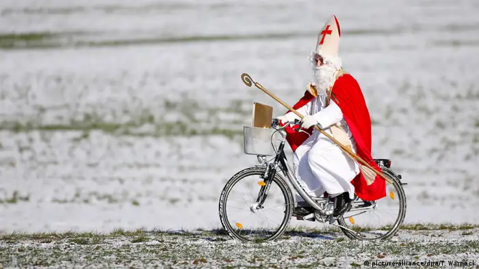 St. Nicholas with staff and mitre rides a bicycle across a snowy meadow, Germany (dpa)