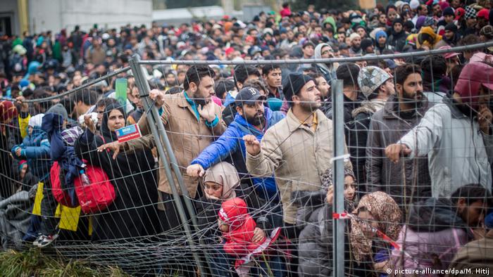 Massen an Flüchtlingen, darunter auch Frauen und Kinder, stehen hinter einem Gitterzaun.