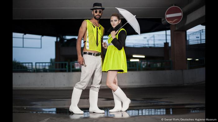Ein Mann und eine Frau posieren in einem Parkhaus; er trägt ein Neon-Shirt, sie ein Neonkleid und einem Regenschirm
