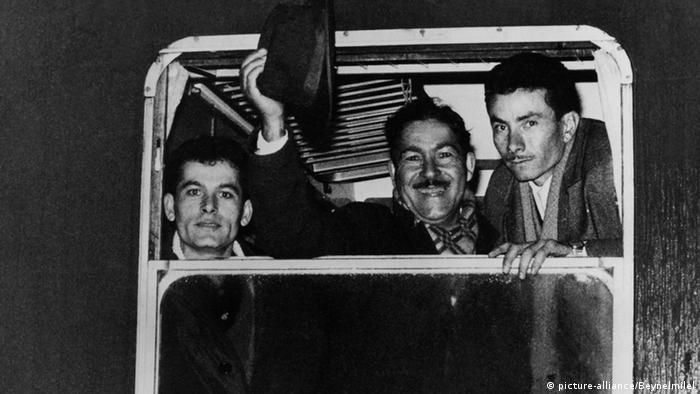 Three Turkish men in the window of a bus, one waving his hat, in 1966 (black and white photo)