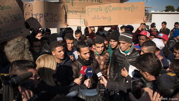 Journalists hold microphones in front of a young man surrounded by other asylum-seekers