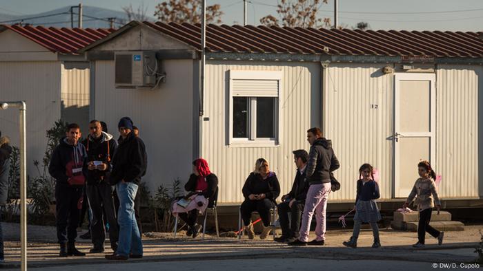 Adults and children stand and sit outside two trailers in Eleonas camp in Athens