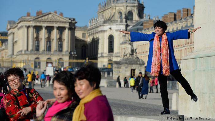 Chinesische Touristinnen posieren für ein Foto vor dem Schloss in Versailles. Eine Frau steht auf dem Podest einer Statue und posiert mit Armen und Beinen abgestreckt. (Foto: Getty Images/P. Le Segretain)