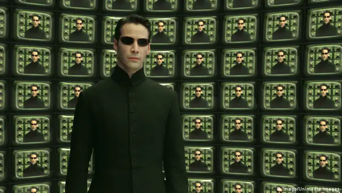 Neo (Keanu Reaves) steht in einer Matrix-Szene mit schwarzer Sonnenbrille und schwarzem Mantel vor unzähligen Bildschirmen, die sein Abbild zeigen. (Imago/Unimedia Images)