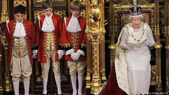 Queen Elizabeth II sits on her throne while wearing her crown with boys standing nearby Queen Elizabeth II sits on her throne while wearing her crown with boys standing nearby