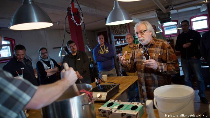 A man holding a beer explains something to a group of people watching as another man mixes something in a big metal pot