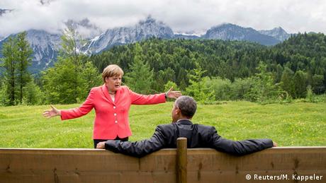 Barack Obama sitzt auf einer Holzbank. Vor ihm steht Angela Merkel und gestikuliert.