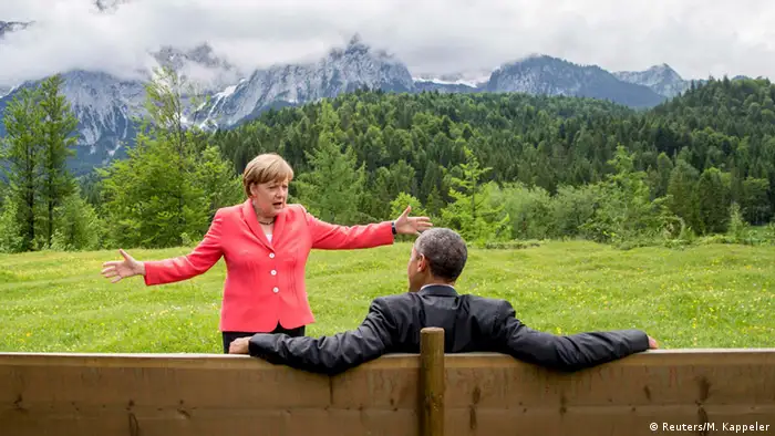 Barack Obama sitzt auf einer Holzbank. Vor ihm steht Angela Merkel und gestikuliert.