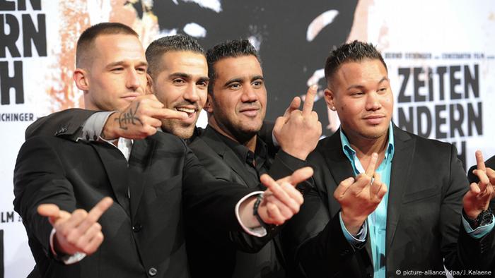 Four men smiling and giving photographers the finger, standing in front of a movie poster