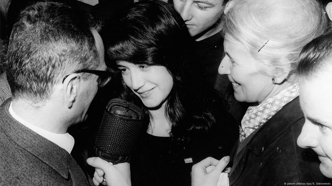 Martha Argerich with long black hair is surrounded by people and readies to speak into a microphone