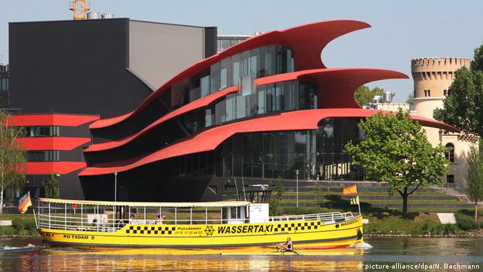 A water taxi passes in front of a futuristic building with red wing-like structures.
