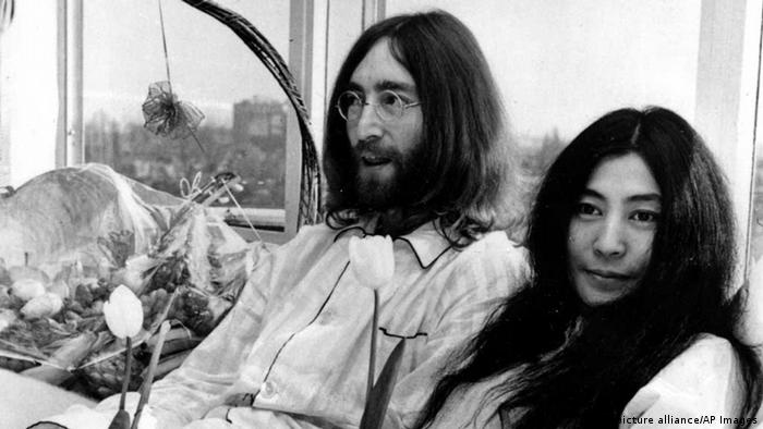Yoko Ono und John Lennen sitzen nebeneinander in einem Bett und halten Tulpen in den Händen.