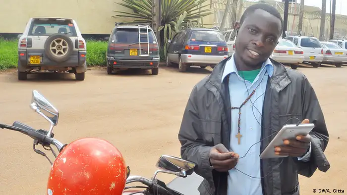 Motorbike taxi driver Mwanje Bernard holds a smartphone, standing in front of his motorcycle.