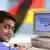 An Indian man sitting in front of a computer with the German flag in the background
