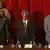 Drei Männer in dunklen Anzügen stehen nebeneinander hinter einem Tisch vor einem roten Vorhang (Quelle: AP)