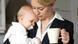 Eine Frau im Hosenanzug hält in einer Hand eine Tasse Kaffee, in der anderen ein Baby Eine Frau im Hosenanzug hält in einer Hand eine Tasse Kaffee, in der anderen ein Baby