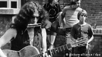 Ein Hippie spielt in den 1970er Jahren Gitarre, im Hintergrund hören drei Jugendliche zu