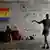 Duas pessoas retratadas num apartamento vazio, com paredes e chão desgastados, entre malas, sacolas e uma bandeira do movimento LGBTI+