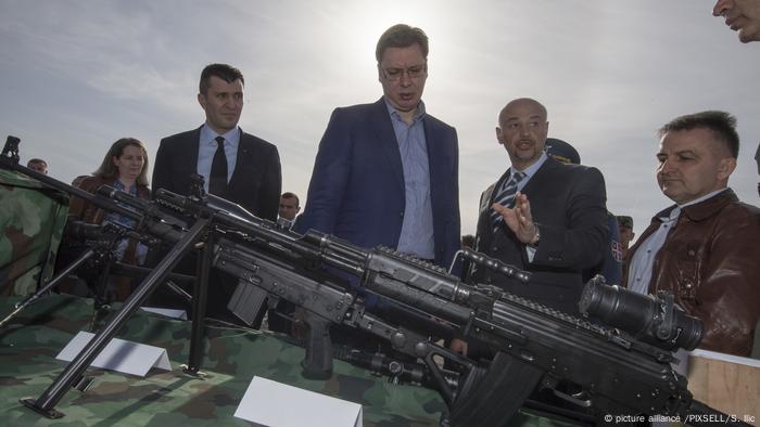 A group of man stand around military weapons, which are in the foreground