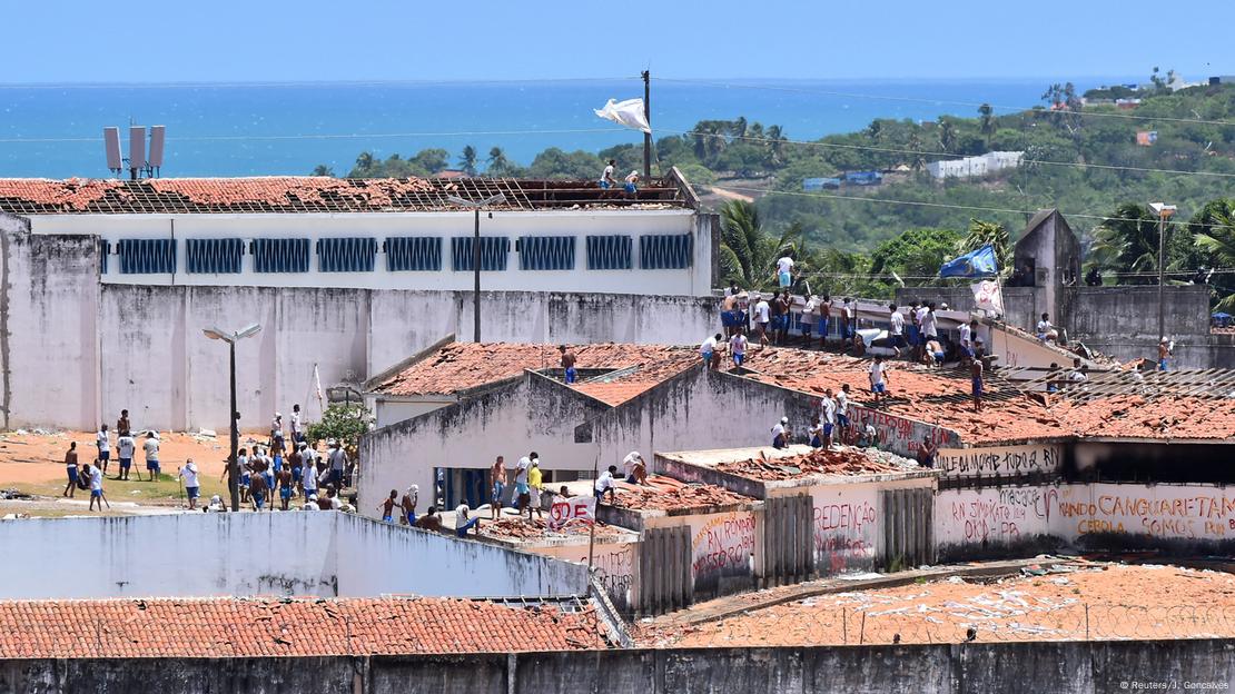 Imagem de Presídio de Alcaçuz, na cidade de Natal, no Rio Grande do Norte. Imagem de 2017 mostra presos em cima do telhado durante rebelião, com mar ao fundo. 