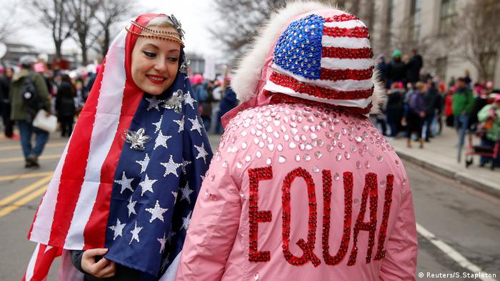 A woman wearing a US flag headscarf gazes at another wearing a pink parka emblazoned with 'Equality' and a US flag hood
