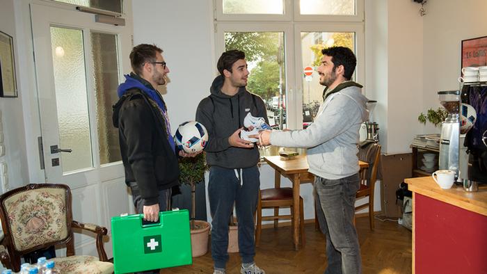 Drei Männer unterhalten sich und haben dabei einen Erste-Hilfe-Koffer, Fußballschuhe und einen Fußball in der Hand. (DW)
