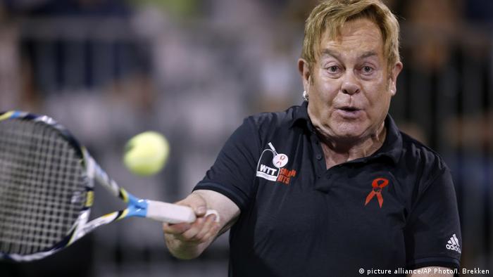 Elton John swings a tennis racket at a tennis ball 