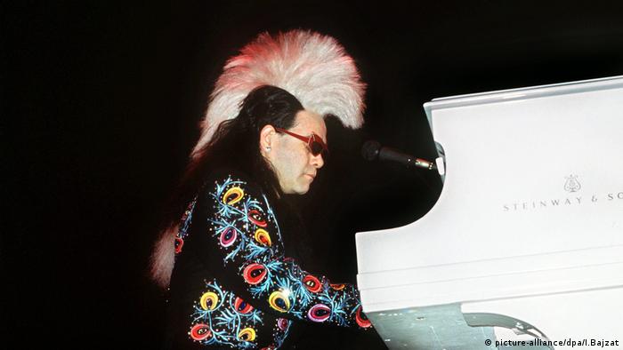 Elton John wearing a wig and colorful jacket at the piano.