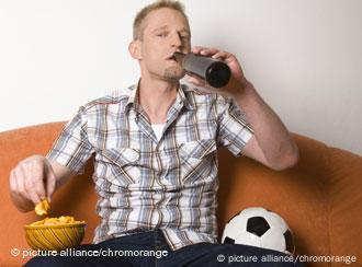 Blonder Mann sitzt mit Chips, Bier und Fußball auf dem Sofa. Quelle: picture alliance/chromorange