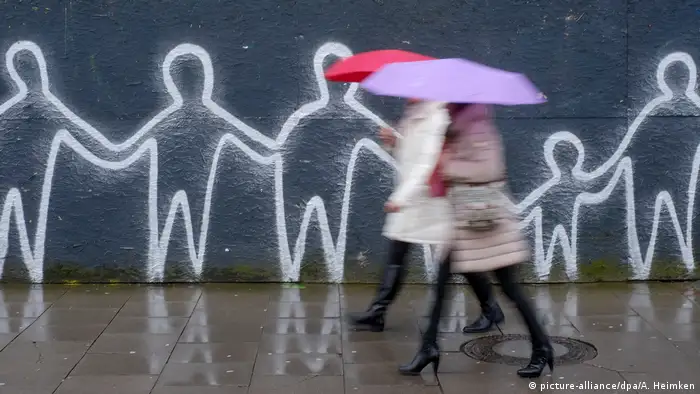 Frauen in Daunenjacken und Regenschirm laufen die Straße entlang, im Hintergrund ist ein Graffiti mit Strichmännchen zu sehen, die sich an der Hand halten (Foto: picture-alliance/dpa/A. Heimken)