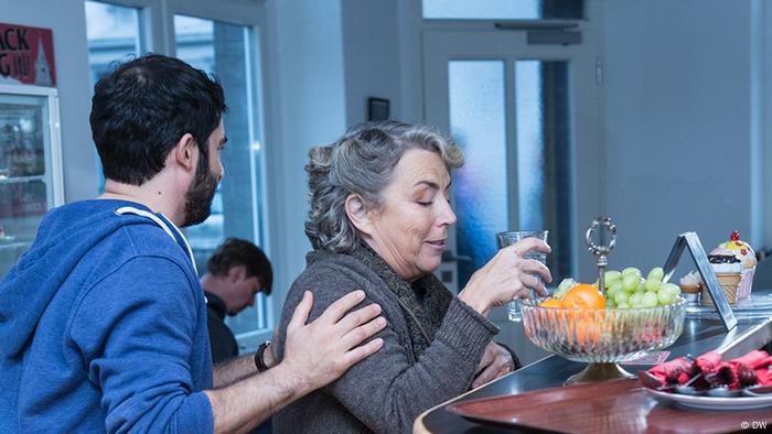 Eine ältere Frau sitzt in einem Café an der Bar und trinkt ein Glas Wasser. Ein jüngerer Mann steht hinter ihr und legt ihr die Hand auf die Schulter.