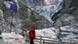 A man steps off a wooden bridge in front of steep, rocky mountain slopes covered with glacial ice and debris A man steps off a wooden bridge in front of steep, rocky mountain slopes covered with glacial ice and debris