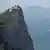 Vista aérea de enorme rochedo pontudo, com mar ao fundo e horizonte com montanhas