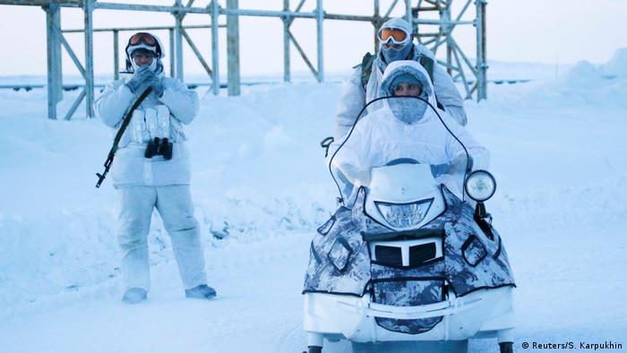 Two Russian servicemen on a snowmobile in all white fatigues. A third serviceman off standing off to the side with a rifle slung on his back. 