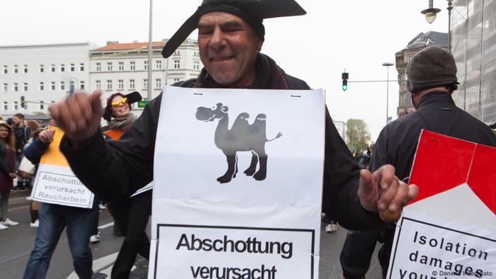 Die Gruppe Tarantella beim Karneval der Kulturen in Berlin 2016. Ein Mann trägt ein weißes Banner mir schwarzer Aufschrift Abschottung verursacht. Darüber ist ein Kamel abgebildet. (Foto: Daniela Incoronato)