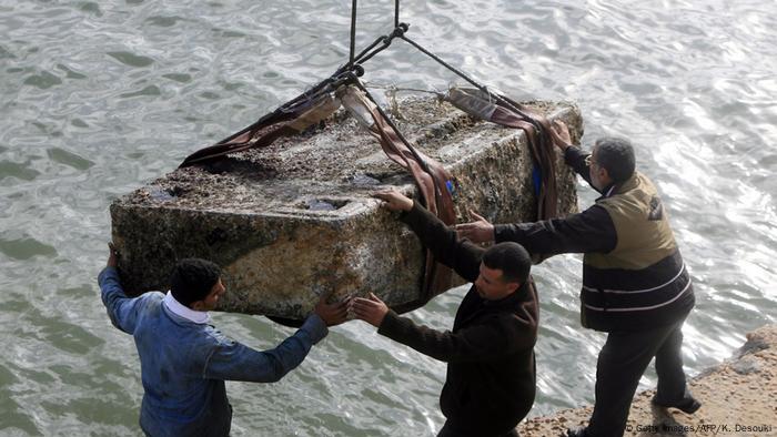 Ein großer Granitblock wird mit einem Kran aus dem Wasser geborgen, drei Männer dirigieren die Bergung