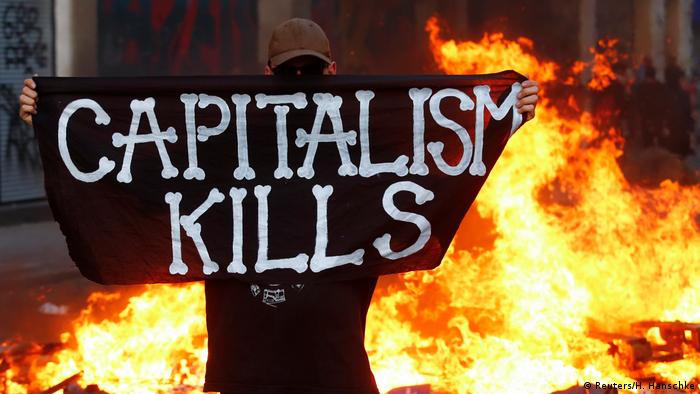 Человек в маске держит плакат с надписью капитализм убивает на фоне горящей баррикады