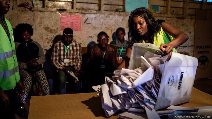 An election commission official tips a ballot box onto a table watched by election monitors