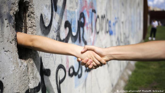 Durch ein Loch im Rest der Berliner Mauer schütteln zwei Personen ihre Hände