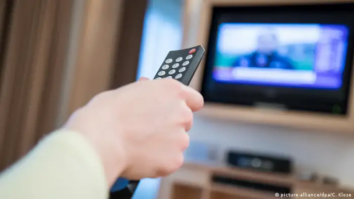 A hand on a TV remote, screen in the background (picture-alliance/dpa/C. Klose)
