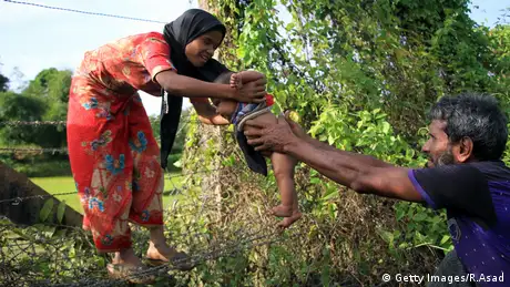 A Rohingya man passes a child though a barbed wire border fence