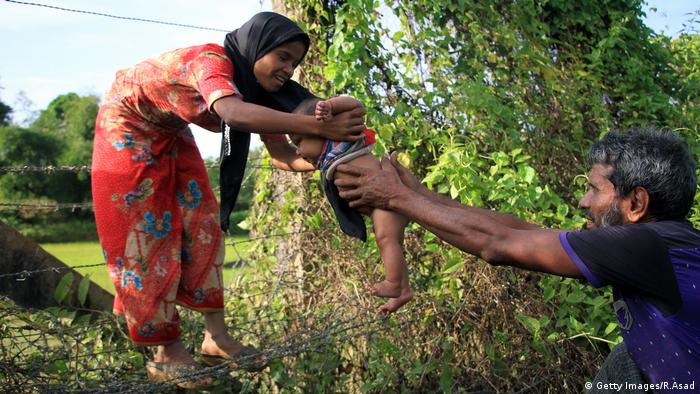 A Rohingya man passes a child though a barbed wire border fence