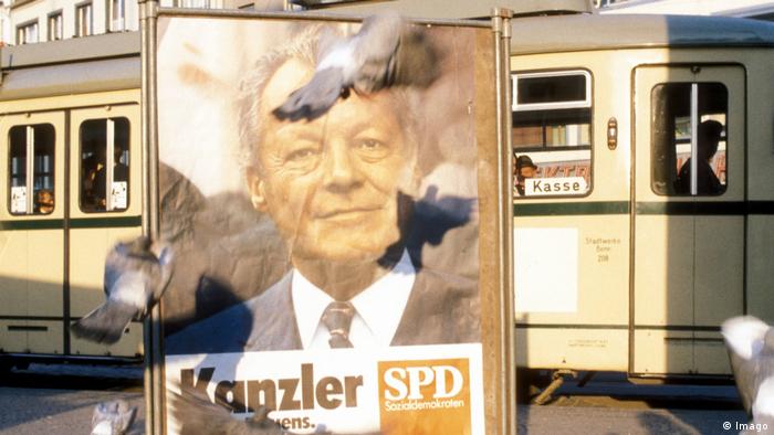 Ein Wahlplakat, aufgehängt ein einer deutschen Innenstadt, zeigt Willy Brandt 1972. Im Hintergrund sieht man eine Straßenbahn. 