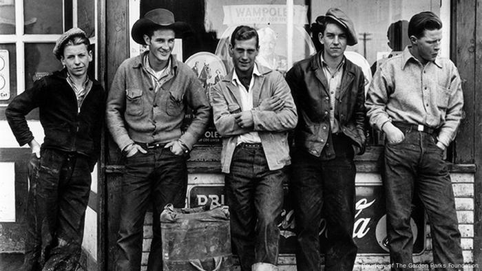 Fotografie von Gordon Parks, Drugstore Cowboys, Turner Valley, Canada, 1945: Fünf junge Männer stehen vor einem Laden und lächeln in die Kamera. Einer schaut zur Seite. (Courtesy of The Gordon Parks Foundation)