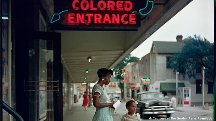Fotografie von Gordon Parks, Department Store, Mobile, Alabama, 1956. Eine schwarze Frau und ein Kind stehen in einer Ladenzeile unter einer Leuchtschrift Colored Entrance (Courtesy of The Gordon Parks Foundation)