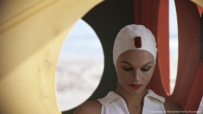 Fotografie von Gordon Parks, Jeweled Cap, Malibu, California, 1958. Eine junge Frau mit weißer Badekappe, aud der ein roter Stein prangt. Sie blickt zu Boden. (Courtesy of The Gordon Parks Foundation)