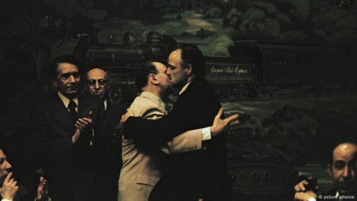Two men hugging, one holding a cigar, with men clapping the background. 